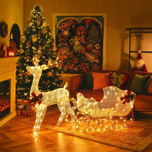 Load image into Gallery viewer, A decoration featuring a lighted reindeer and Santa's sleigh with Christmas trees and presents in the background, on a room with a fireplace.