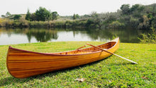 Load image into Gallery viewer, A wooden canoe with ribs and a curved bow placed on a grassy area near a body of water, accompanied by a wooden paddle.